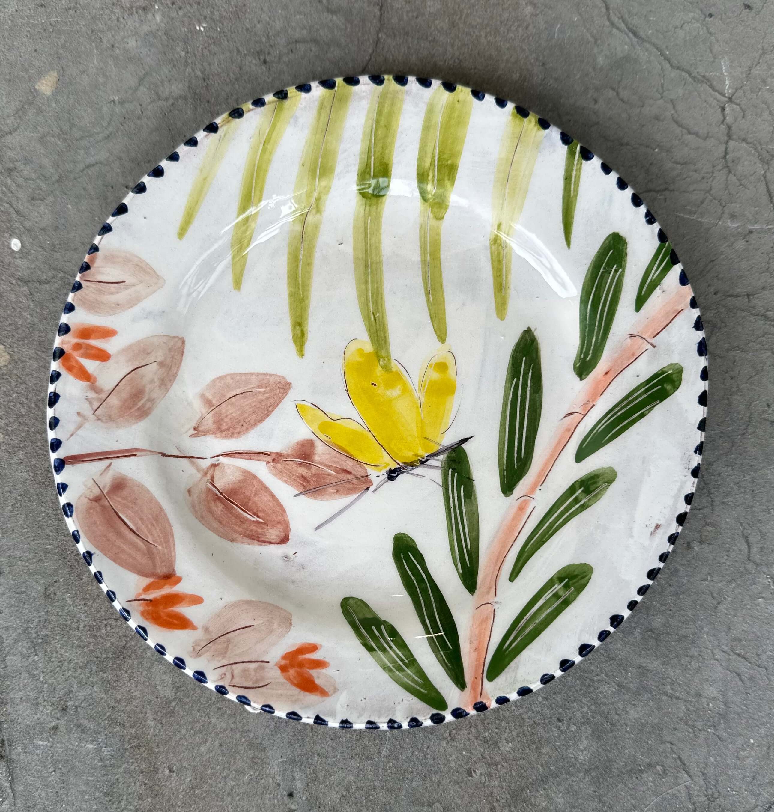 A handmade ceramic plate with a yellow butterfly design surrounded by green leaves and a blue rim on a textured gray background.