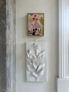 'Votive IV' by Becky Blair pictured on a white wall above a plaster relief